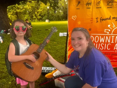 A young child trying a guitar with a Downingtown Music Academy teacher at the studio's booth at an outdoor community event